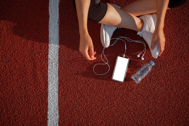 Trening i fitness na stadionie. Dziewczyna biegacza trzyma smartfon za pomocą ekranu dotykowego, aby wybrać muzykę lub tekst w aplikacji przed uruchomieniem na torze.