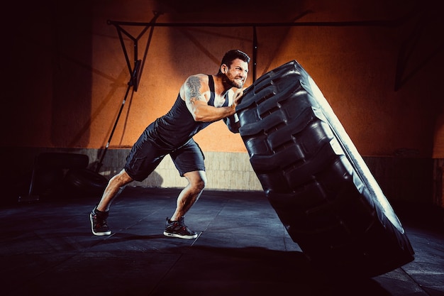 Trening kulturystyczny, brodaty silny sportowiec z umięśnionym ciałem podnoszącym ciężkie koło na siłowni.