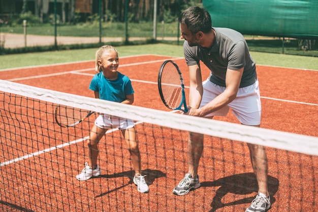 Trening tenisowy. Wesoły ojciec w sportowej odzieży uczy córkę grać w tenisa, gdy oboje stoją na korcie tenisowym