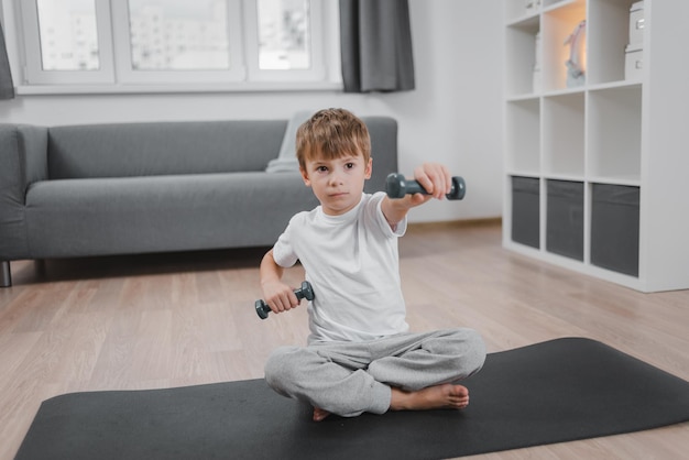 Trening w domu. Portret chłopca, ćwiczenia z hantlami, siedząc na podłodze w salonie.