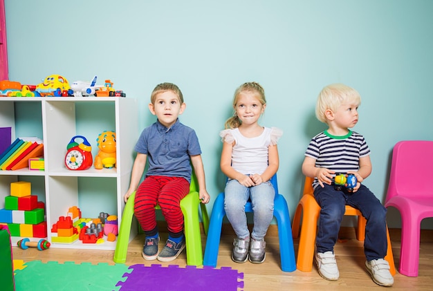 Troje dzieci w wieku przedszkolnym siedzi na kolorowych plastikowych krzesełkach pod ścianą w przedszkolu patrząc na coś z dużym zainteresowaniem Dzieci w przedszkolu podczas imprezy