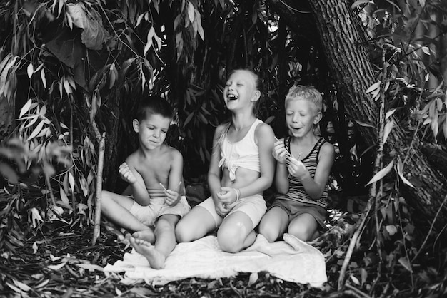 Troje wiejskich dzieci bawi się w chacie, którą same zbudowały z liści i gałązek Drewniany dom w lesie Fotografia czarno-biała