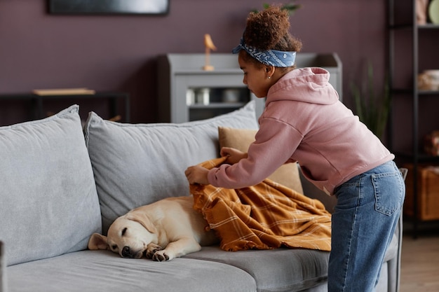 Troskliwa czarna dziewczyna kładąca koc nad uroczym psem śpiącym na kanapie w domu