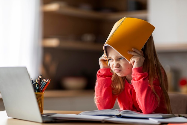 Zdjęcie trudności w nauce irytowały małą dziewczynkę zakrywającą głowę zeszytem ćwiczeń w domu