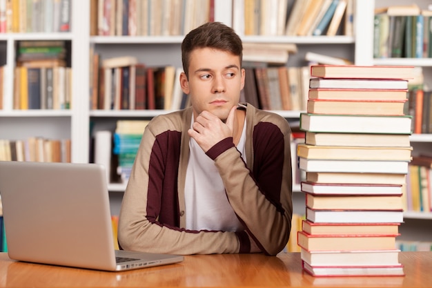 Trudny Wybór. Zdezorientowany Młody Mężczyzna Siedzący Przy Biurku W Bibliotece Ze Stosem Książek I Laptopem Na Nim
