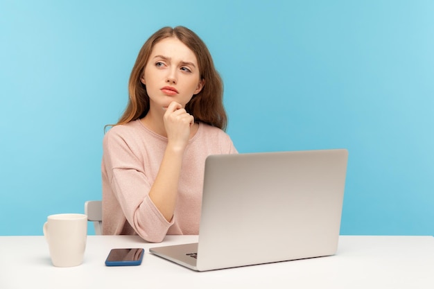 Trzeba przemyśleć biznesplan Zamyślona kobieta w zwykłych ubraniach siedzi z zamyślonym zdezorientowanym wyrazem twarzy podczas pracy na laptopie w domowym biurze wewnątrz studio strzał na białym tle na niebieskim tle