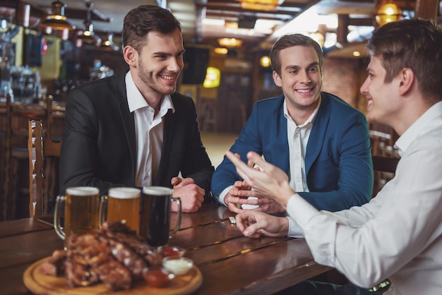 Trzech młodych biznesmenów w garniturach uśmiecha się, rozmawiając i pijąc piwo siedząc w pubie