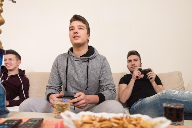 Trzech młodych graczy siedzących razem na kanapie i grających w gry wideo w domu