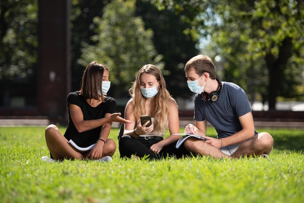 Trzech studentów siedzi i rozmawia w parku w masce na twarz