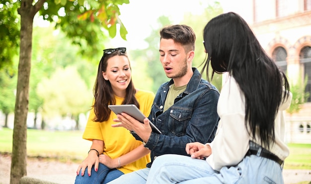 Trzech studentów uczących się razem z cyfrowym tabletem siedząc na ławce na świeżym powietrzu