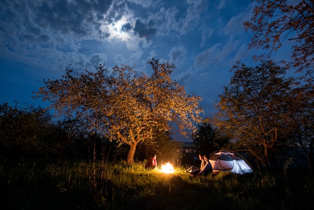 Trzech turystów siedzących przy ognisku w pobliżu namiotu pod drzewami i nocnym niebem z księżycem. Nocne biwakowanie