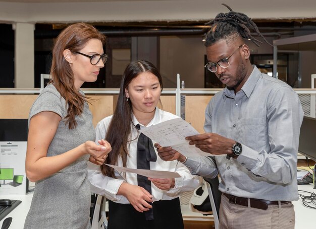 Trzech wieloetnicznych partnerów biznesowych omawiających papierkową robotę w biurze, trzymających dokumenty i rozmawiających