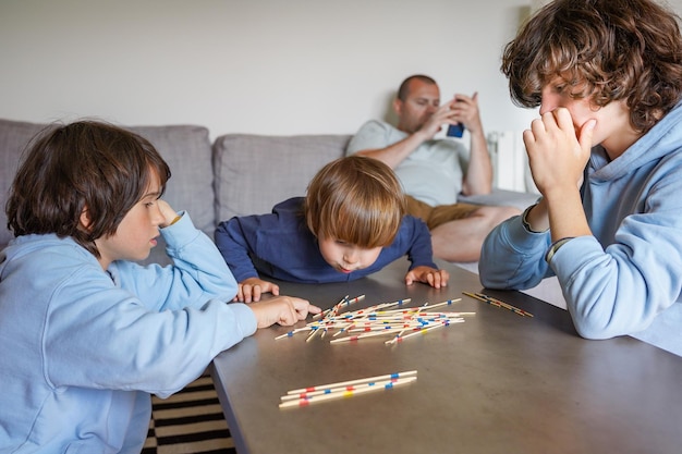 Zdjęcie trzej bracia grają w domu w grę planszową mikado, podczas gdy ojciec sprawdza na smartfonie, zabawne rodzeństwo spędzające czas razem