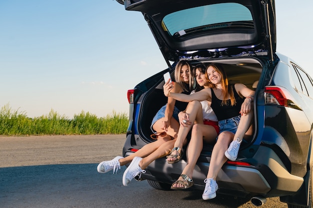 Trzy dziewczyny robią sobie razem selfie. Blondynka, brunetka i rudowłose młode kobiety usiąść i zrelaksować się z tyłu pojazdu, rozmawiając podczas podróży przygodowej w przyrodzie.