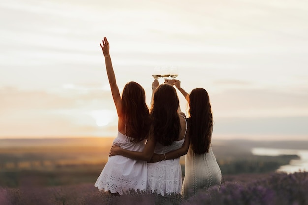 Trzy kobiety w białych sukienkach trzymają kieliszek wina przed zachodem słońca