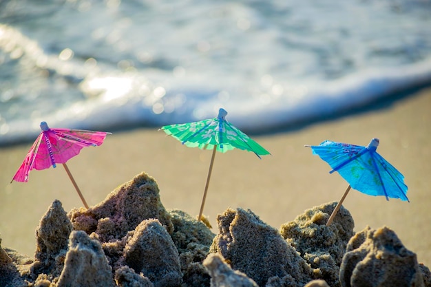 Trzy małe papierowe parasole plażowe na stojak na koktajle w piasku na piaszczystej plaży nad brzegiem morza lub oceanu i toczące się fale w słoneczny dzień zbliżenie Koncepcja szczęście radość relaks turystyka wakacje podróże