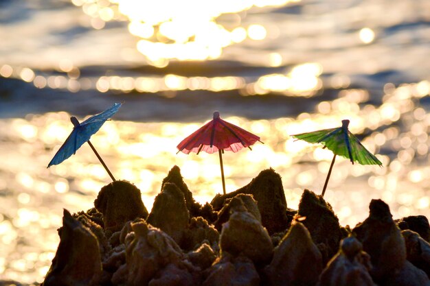 Trzy małe parasole plażowe wykonane z papieru do stoiska koktajlowego w piasku na piaszczystej plaży brzegu morza lub oceanu z rozmytym odblaskiem słońca na powierzchni fal z bliska. Koncepcja szczęście radość relaks