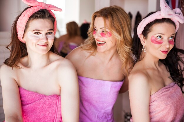 Trzy piękne młode kobiety w różowych ręcznikach, z kosmetycznymi bandażami na głowach, pozują do kamery z łatami pod oczami. .