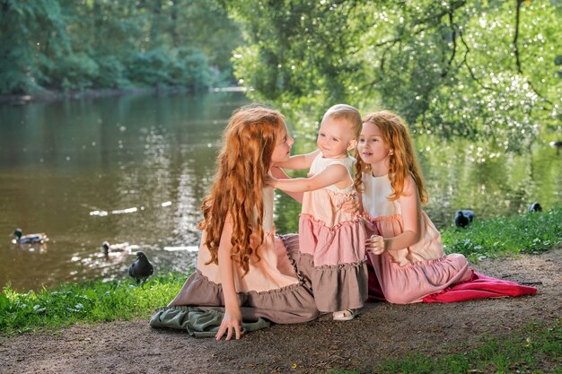 Trzy rudowłose siostry w długich lnianych sukienkach dmuchają w parku w słoneczny letni dzień.
