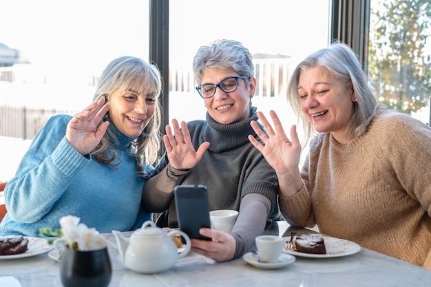 Trzy starsze przyjaciółki korzystające ze smartfona i sieci społecznościowej do wysyłania wiadomości wideo i prowadzenia rozmów wideo w kafejce stylu życia dojrzałych kobiet i szybkim momencie