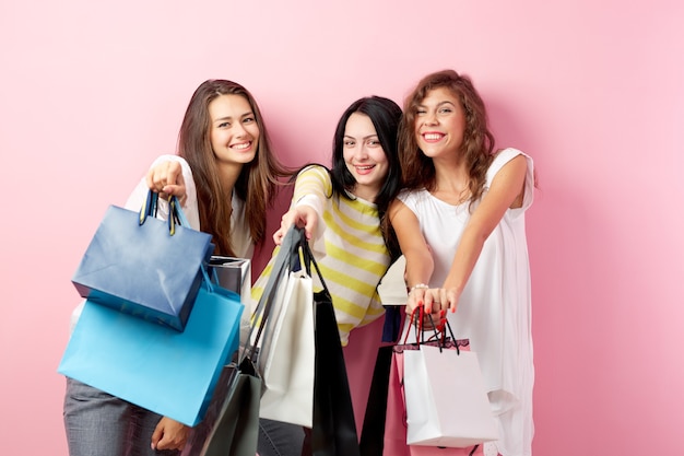 Trzy szczęśliwe dziewczyny ubrane w ładne stylowe ubrania stoją z mnóstwem toreb po zakupach na różowym tle.