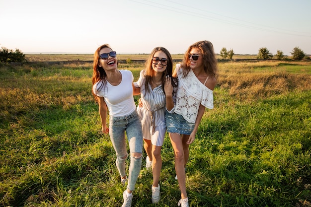 Trzy wspaniałe młode kobiety w okularach przeciwsłonecznych, ubranych w piękne ubrania stoją w polu i uśmiechają się w słoneczny dzień. .
