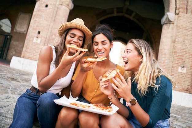 Trzy zabawne kobiety z różnych ras jedzące pizzę na ulicy, szczęśliwe przyjaciółki cieszące się