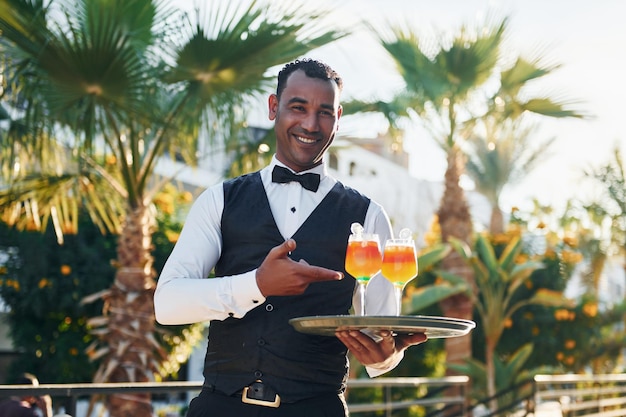 Trzyma koktajle Czarny kelner w eleganckim stroju pracuje na dworze w słoneczny dzień