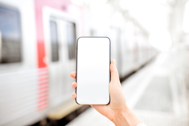 Trzymając smartfona z pustym ekranem z pociągiem na tle na stacji kolejowej