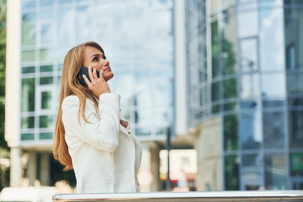 Trzymając telefon w dłoni Kobieta w stroju wizytowym stojąca na zewnątrz w mieście w ciągu dnia