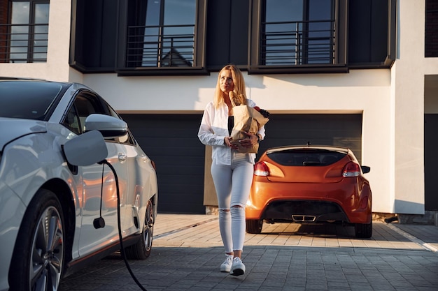 Trzymając torbę na zakupy Młoda kobieta w białych ubraniach jest ze swoim samochodem elektrycznym w ciągu dnia