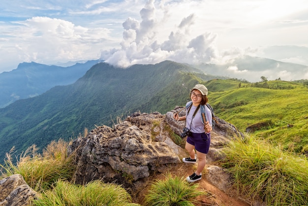 Turysta Asian teen dziewczyna zadowolony z kamery plecak czapka i okulary patrząc piękny krajobraz natura na górze i kolorowe niebo o zachodzie słońca, Travel Asia at Phu Chi Fa Forest Park, Chiang Rai, Tajlandia