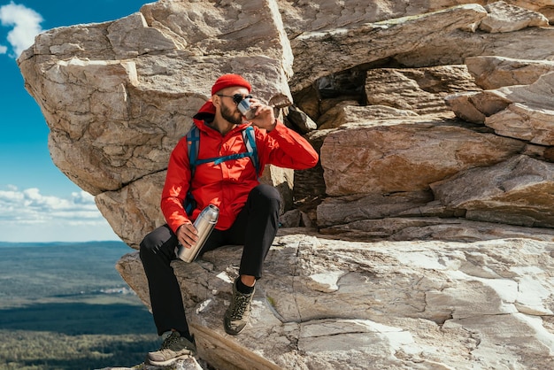 Turysta mężczyzna odpoczywa po ciężkiej wspinaczce na górę. Turysta pije herbatę z termosu w górach. Podróżnik pije gorącą kawę z termosu w górach. Skopiuj miejsce