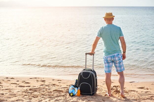 Turysta mężczyzna w letnie ubrania z walizką w ręku, patrząc na morze na plaży.