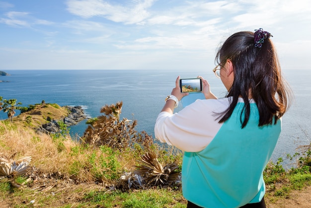 Turystka fotografująca wyspę i morze na wysokim wzgórzu za pomocą smartfona w punkcie widokowym Cape Phromthep, słynne atrakcje prowincji Phuket w Tajlandii