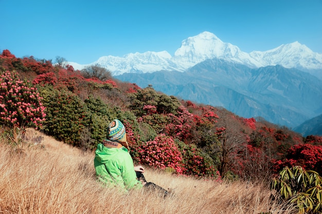 Zdjęcie turystyczna dziewczyna w jasnym nepalskim kapeluszu siedzi na tle ośnieżonych himalajów i kwitnących rododendronów. piękny widok kwiatonośni rododendronowi drzewa i śnieżne góry, nepal