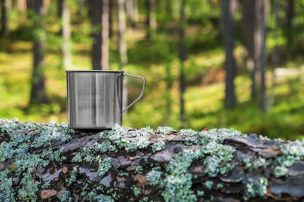 Turystyczny metalowy kubek z gorącym napojem stoi na pniu drzewa w lesie Turystyka na świeżym powietrzu
