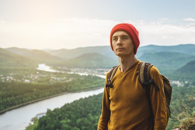 Turystyczny podróżnik z plecakiem i czerwonym kapeluszem stoi na skraju urwiska i patrzy na zieloną dolinę z rzeką