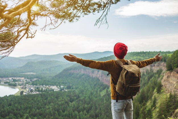 Turystyczny podróżnik z plecakiem i czerwonym kapeluszem stoi na skraju urwiska przed zieloną doliną z szeroko rozłożonymi rękami