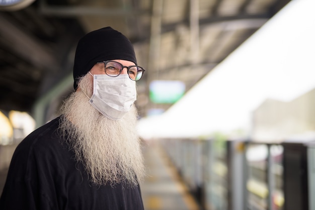 Twarz dojrzałego brodatego mężczyzny myślącego z maską do ochrony przed wybuchem wirusa koronowego na stacji kolejowej