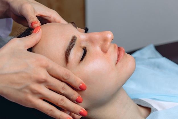 Twarz młodej kobiety z zamkniętymi oczami. Dłonie kosmetologa wykonują jej odmładzający masaż.