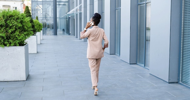 Tył African American Piękna Kobieta W Masce Medycznej Rozmawia Przez Telefon Komórkowy I Chodzenie Na Zewnątrz W Centrum Biznesowym. Szczęśliwy Bizneswoman Mówi Na Telefon Komórkowy I Spacery. Widok Z Tyłu.