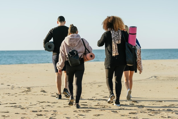 Tył grupy czterech wieloetnicznych ludzi spacerujących po plaży z matami do jogi i torbami