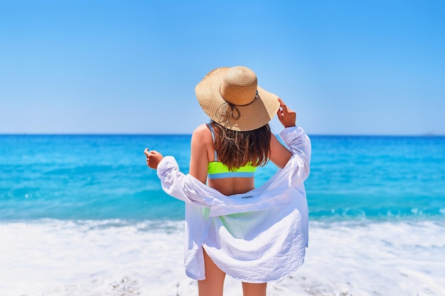 Tył modnej wspaniałej stylowej seksownej szczupłej pięknej brunetki w słomkowym kapeluszu jasny strój kąpielowy i koszula na plaży w letni słoneczny dzień