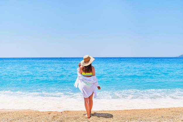 Tył modnej wspaniałej stylowej seksownej szczupłej pięknej brunetki w słomkowym kapeluszu jasny strój kąpielowy i koszula na plaży w letni słoneczny dzień