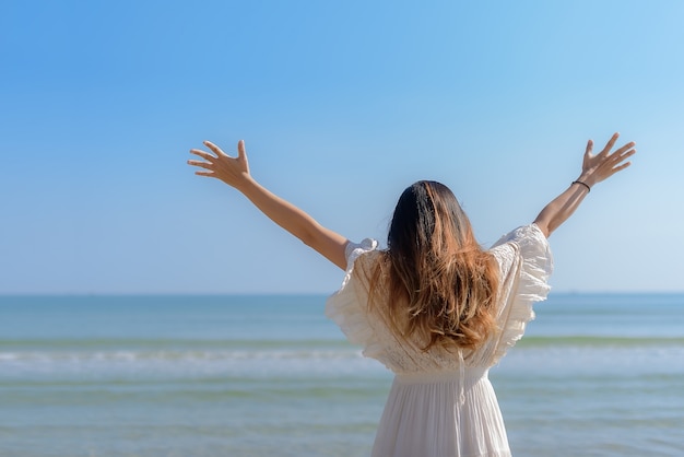 Zdjęcie tył pięknej dziewczyny wyciąga ręce i stoi na plaży pośród błękitnego nieba
