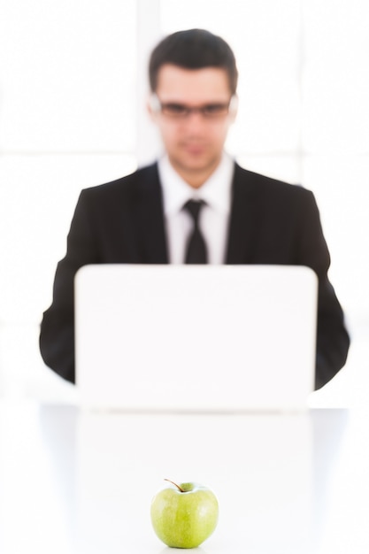 Tylko świeże pomysły. Pewny siebie młody człowiek w formalwear pracujący na komputerze siedząc w swoim miejscu pracy z zielonym jabłkiem leżącym na pierwszym planie