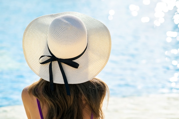 Tylny widok młoda kobieta relaksuje w lecie blisko pływackiego basenu z błękitne wody na słonecznym dniu z długie włosy jest ubranym słomianym kapeluszem.