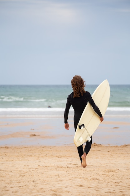 Tylny widok młody człowiek w czarnym kombinezonu odprowadzeniu w kierunku morza z surfboard