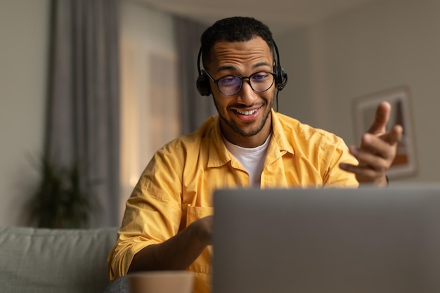Zdjęcie tysiąclecia czarny mężczyzna w zestawie słuchawkowym podczas rozmowy internetowej za pomocą laptopa siedzącego na kanapie rozmawiającego przez kamerę internetową w domu